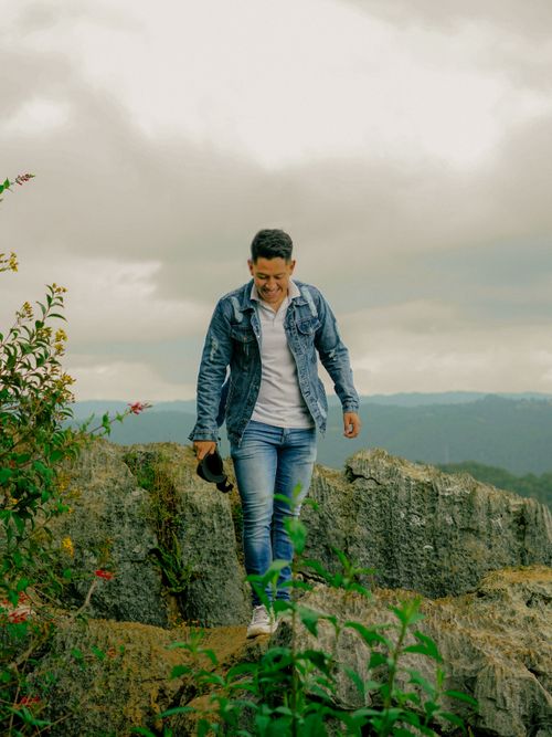 Free Man in denim jacket exploring rocky mountain trail, smiling with nature in background. Stock Photo
