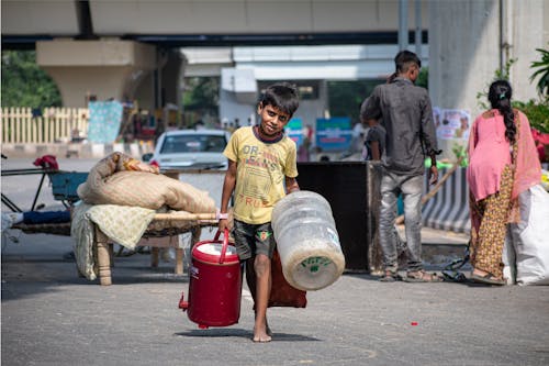 A Boy Holding Containers and Walking Barefoot on a Street 