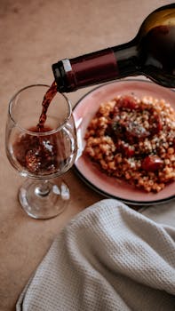 Elegant dining scene with red wine being poured into glass beside a plate of Italian pasta.