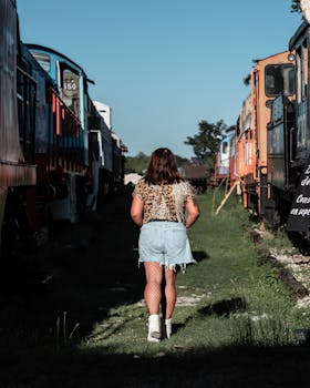 Back view of a woman walking between vintage trains outdoors on a sunny day.
