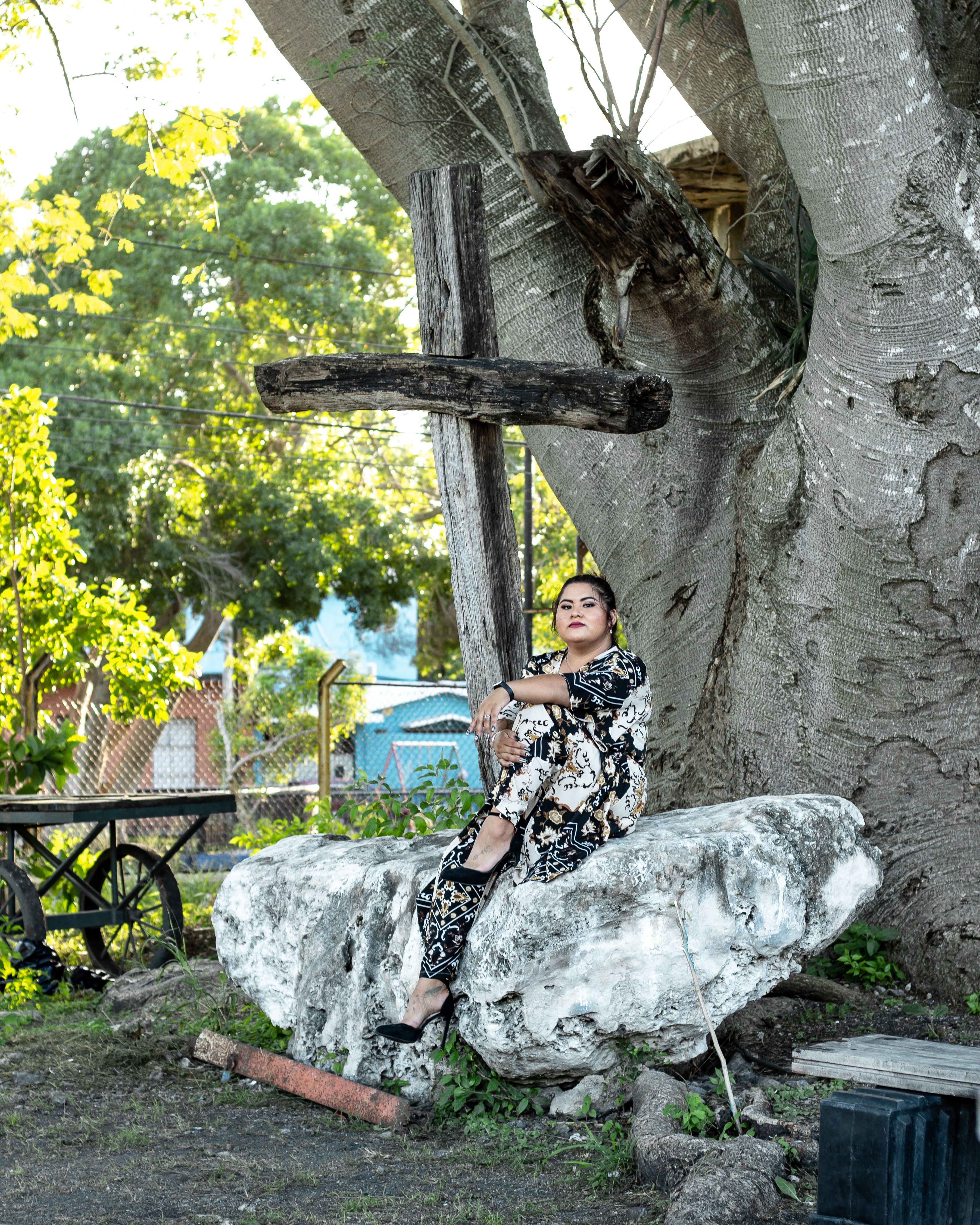 Woman in patterned outfit sitting beside a wooden cross on a large rock outdoors, surrounded by nature.
