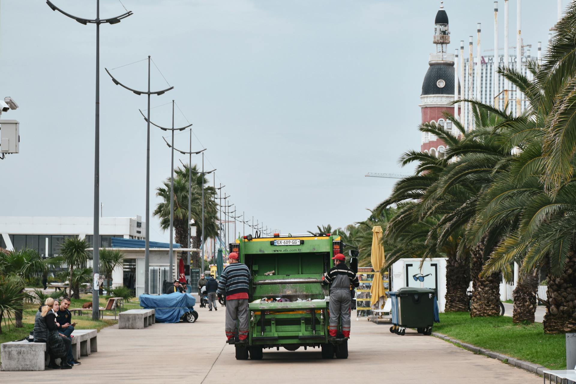 A vibrant urban scene featuring a garbage truck on a street lined with palm trees and people enjoying the promenade.