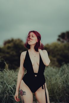 Redhead woman posing in a black swimsuit with a leg tattoo on a cloudy day outdoors.