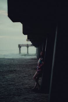 A woman standing under a pier at sunset, casting a serene silhouette on the beach.