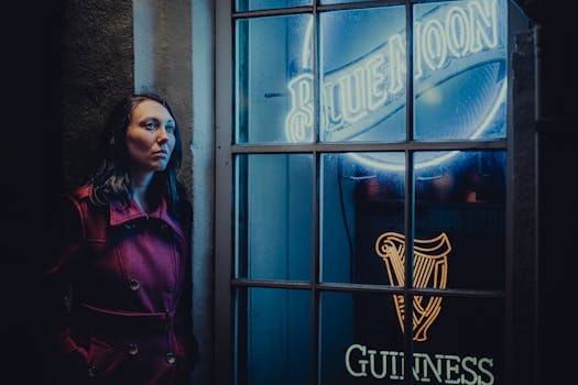 Woman in a red coat leans against a wall by blue neon signs at night.