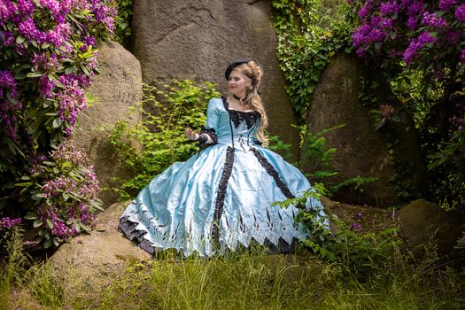 A woman in a vintage blue gown poses amongst flowering shrubs and rocks, evoking historical elegance.