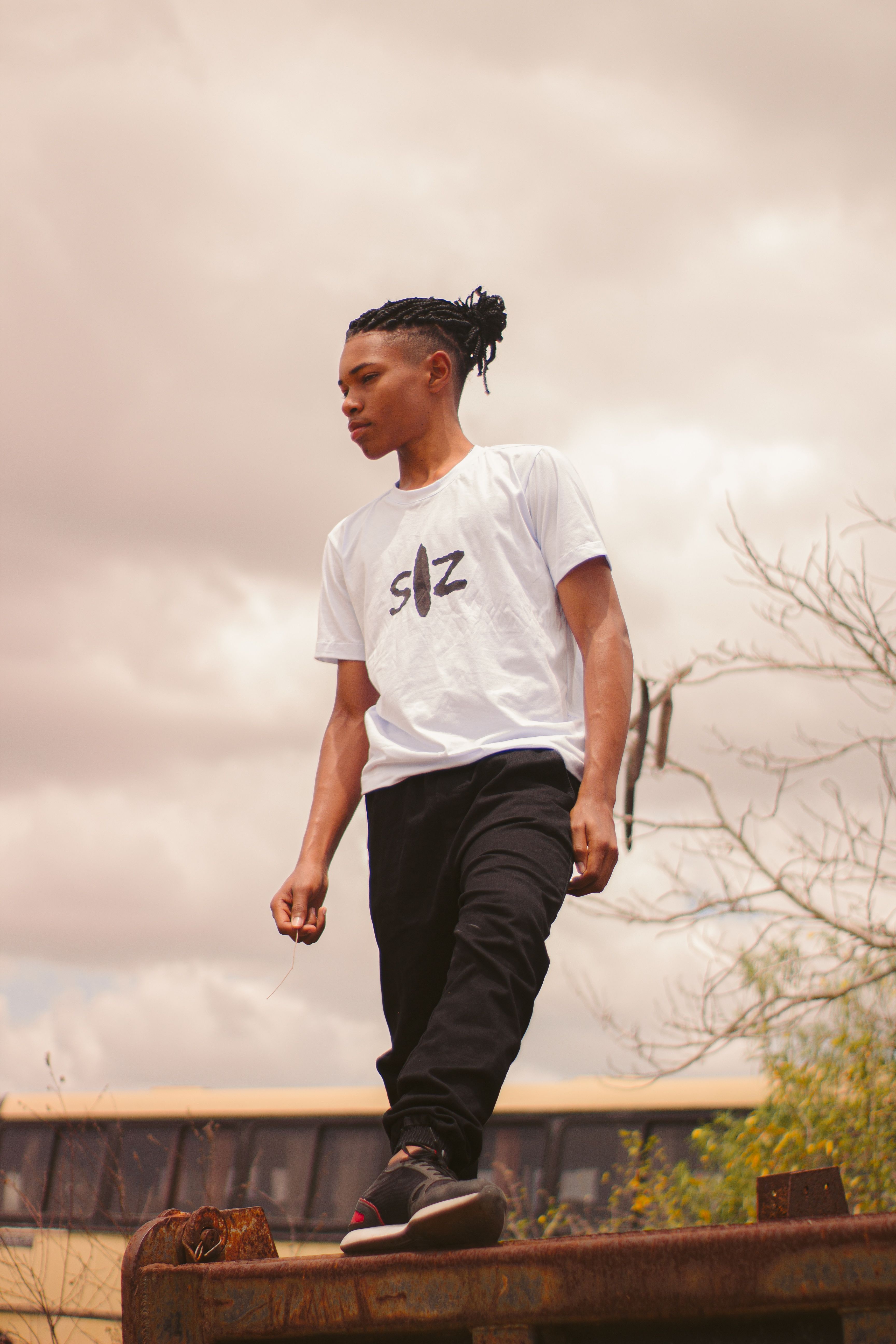 A young man with dreadlocks and a casual outfit poses confidently outdoors under a cloudy sky.