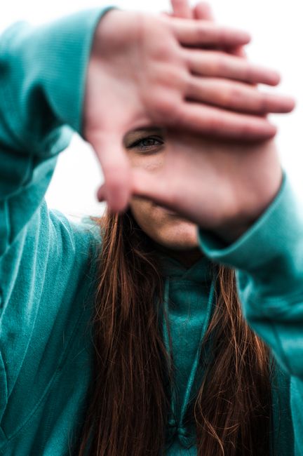 Portrait of a woman peering through her hands creating a frame, wearing a teal hoodie outdoors.