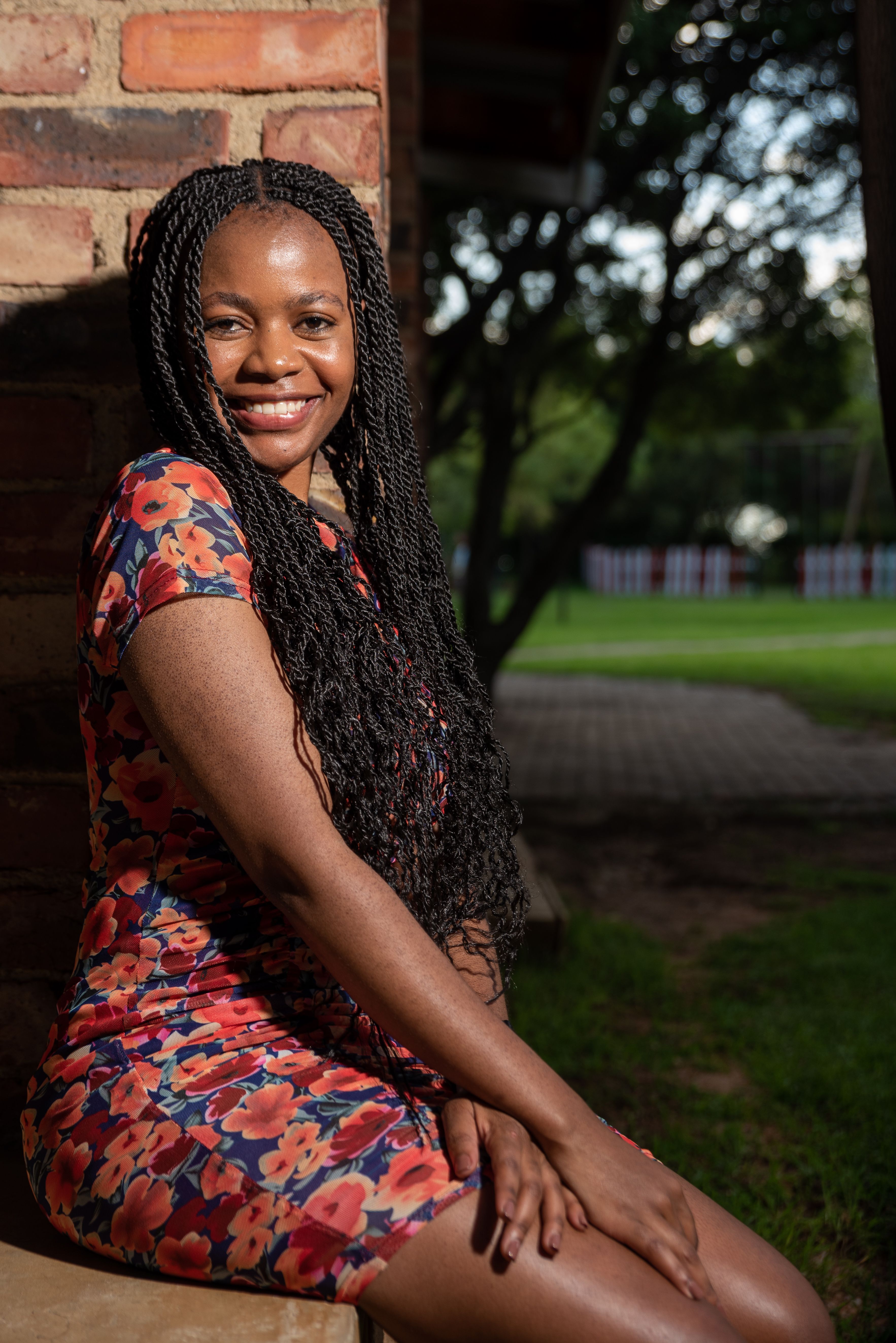 Portrait of a stylish woman with braided hair wearing a floral dress, smiling while sitting outside.