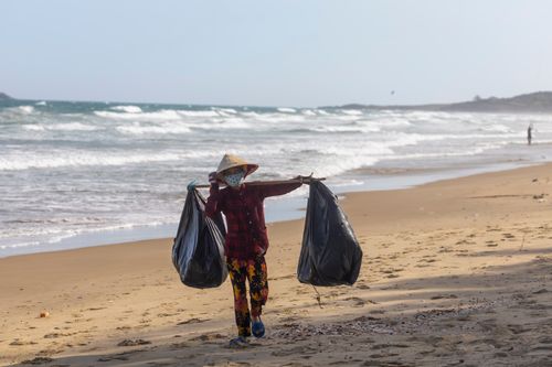 Free Person in a rice hat and facemask collecting trash on a beach, contributing to environmental conservation. Stock Photo