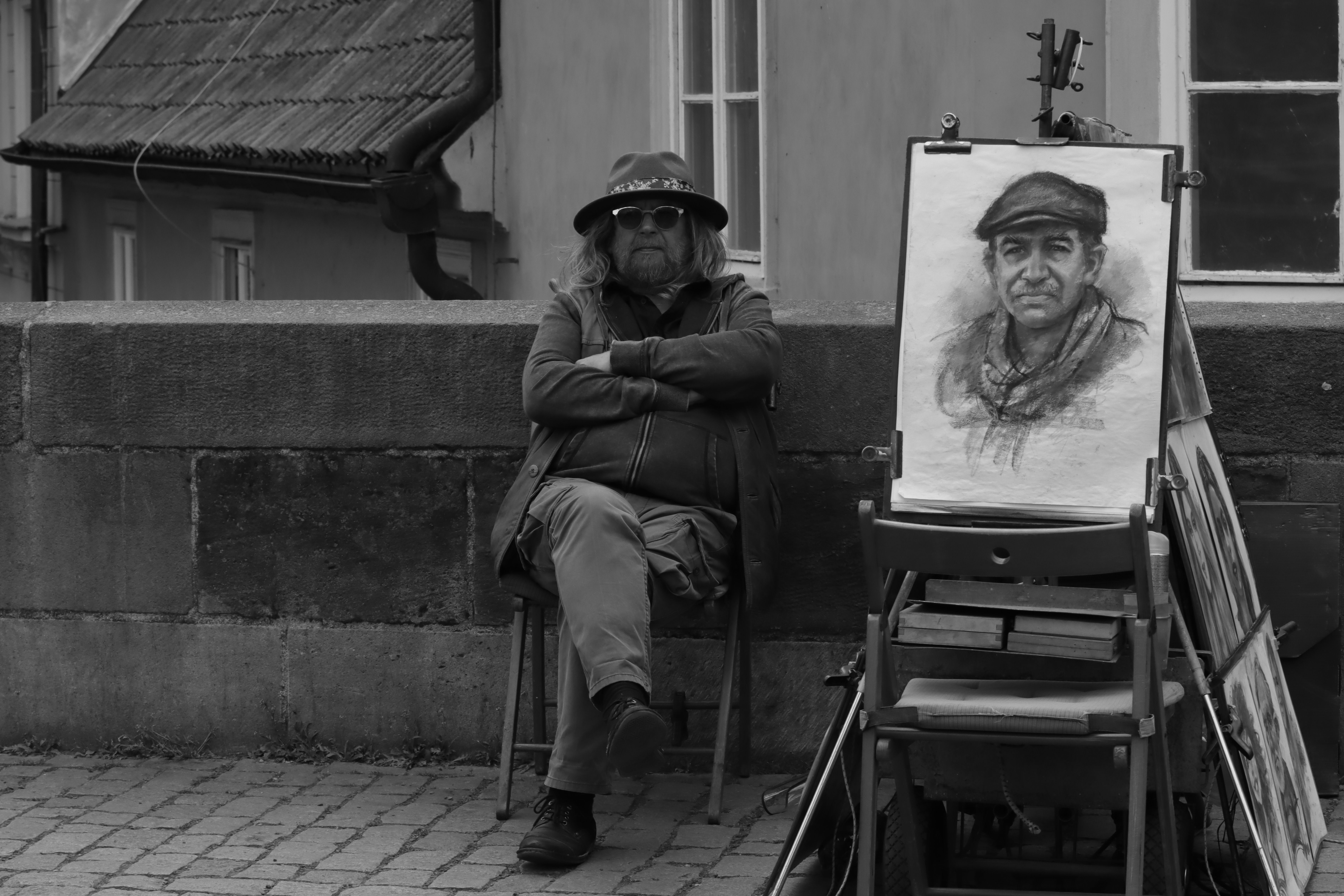 A street artist sits with his portrait sketch on display in a grayscale urban scene.