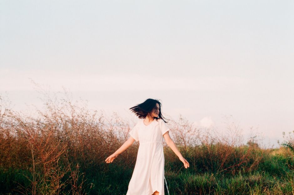 A woman in a white dress with black hair spins freely in a grassy field, capturing a sense of freedom.