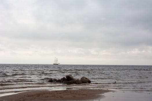 A tranquil scene of a sailboat on the horizon under a cloudy sky, with gentle waves and rocks on the shore.
