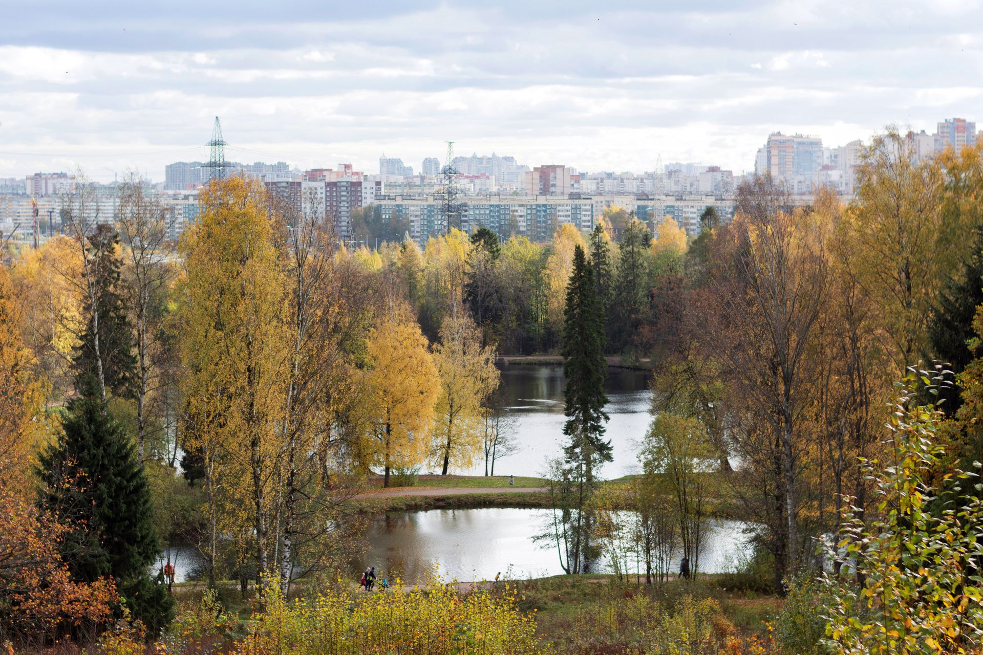 Aerial view of a picturesque park with autumn foliage, a serene lake, and a city skyline in the background.