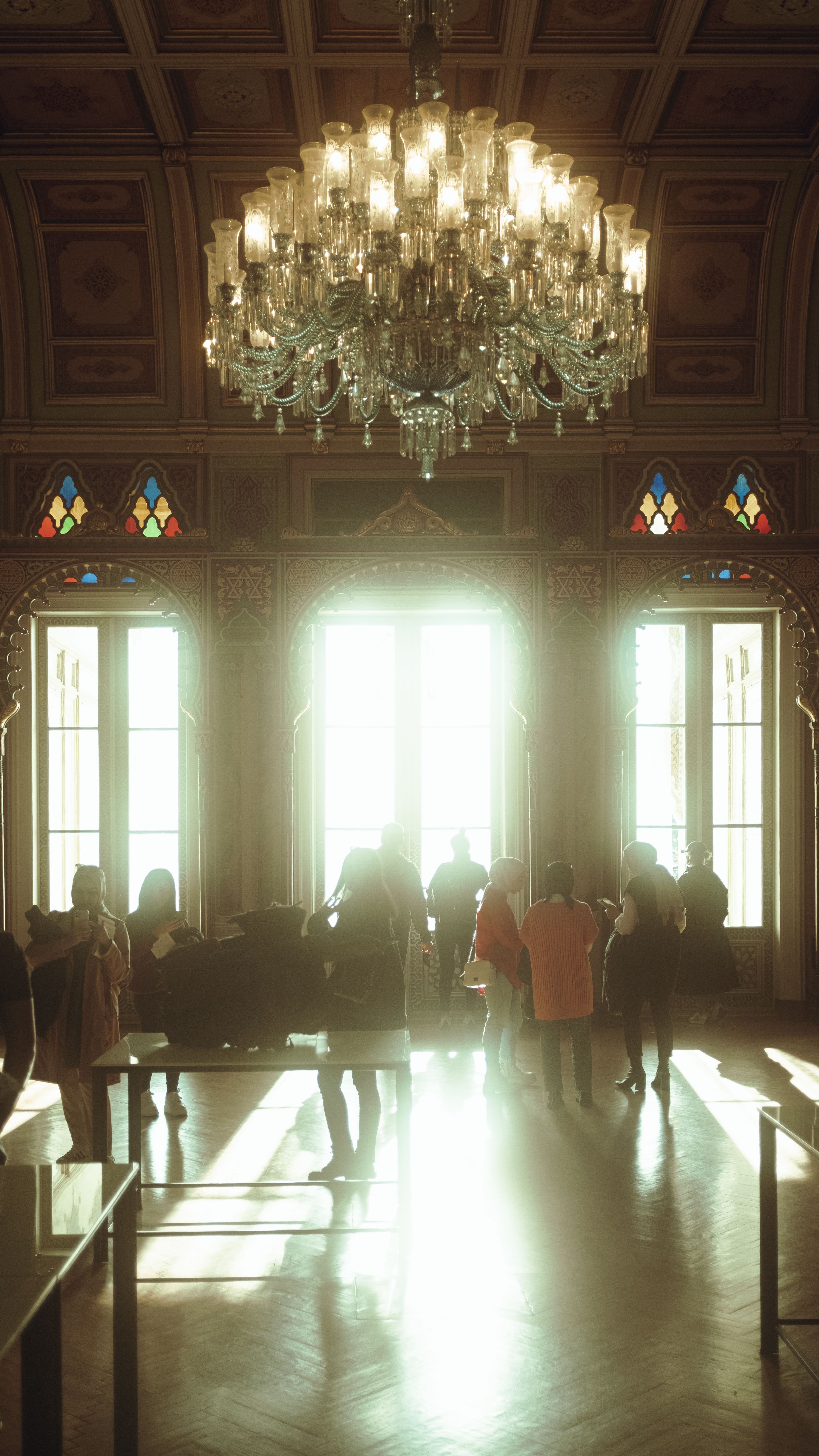 A grand ballroom featuring a majestic chandelier and stained glass windows with people mingling.