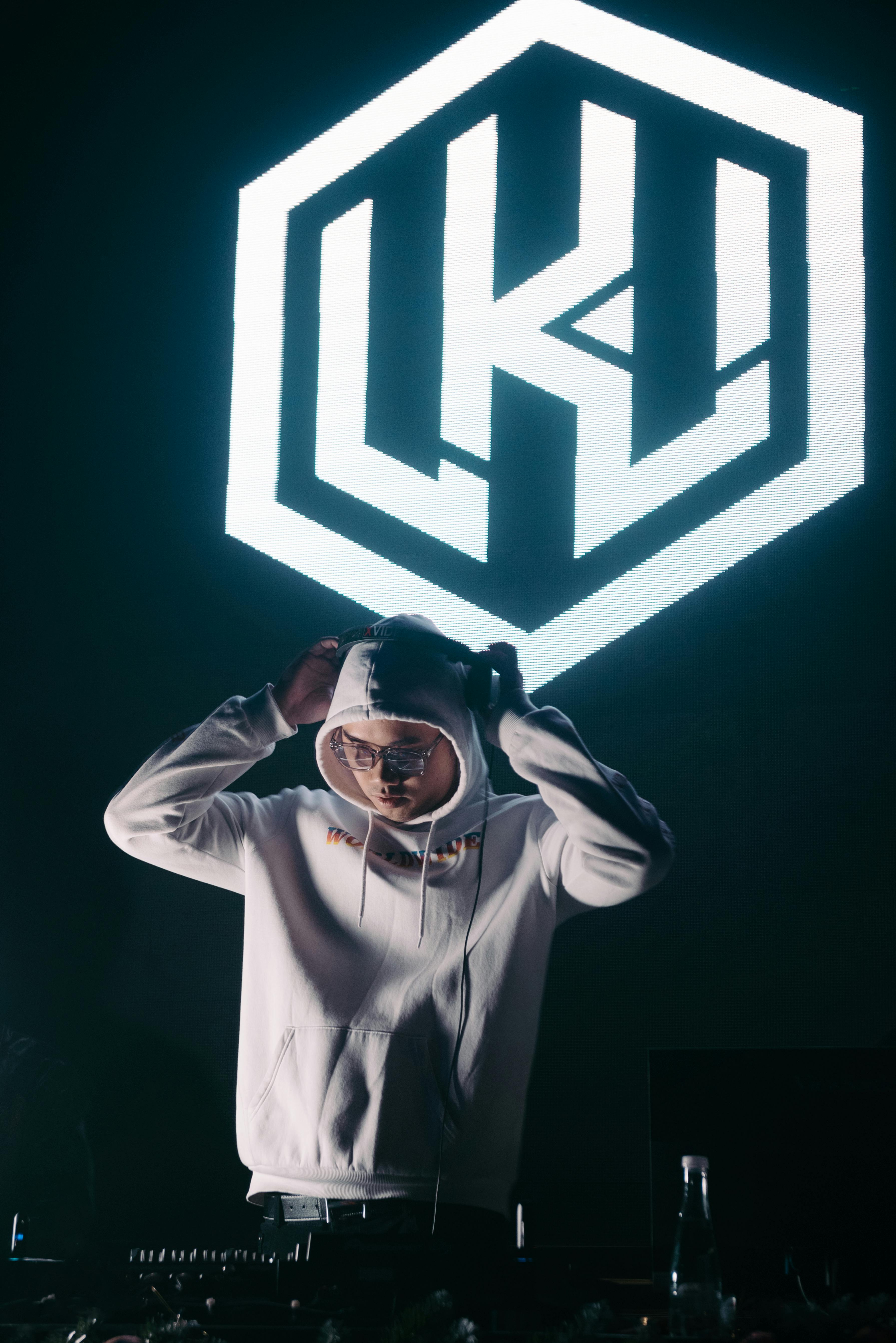 DJ wearing eyeglasses and white hoodie performing under illuminated signage in nightclub.