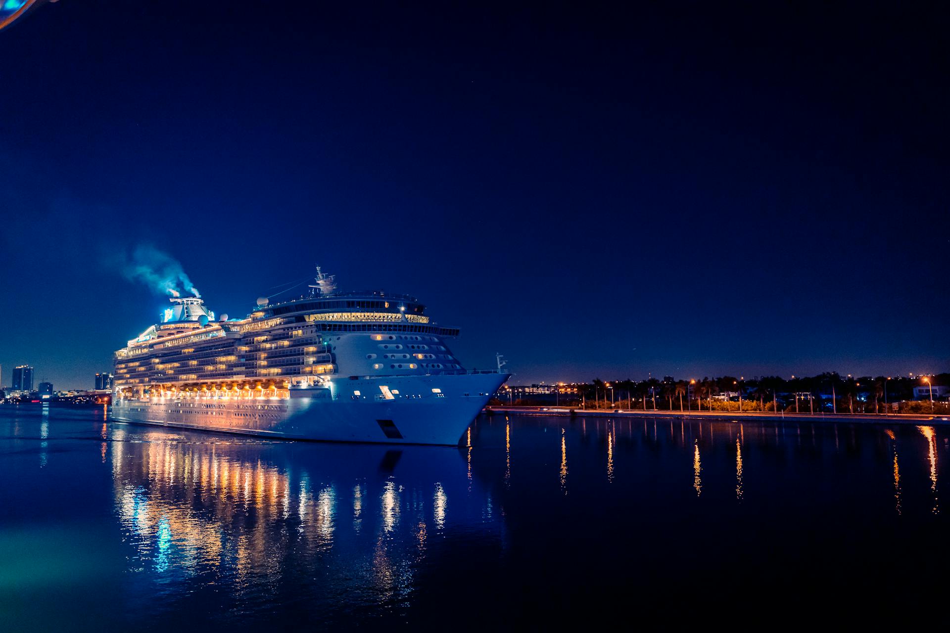 A brightly lit luxury cruise ship sails through Miami's waters at night, reflecting in the calm sea.