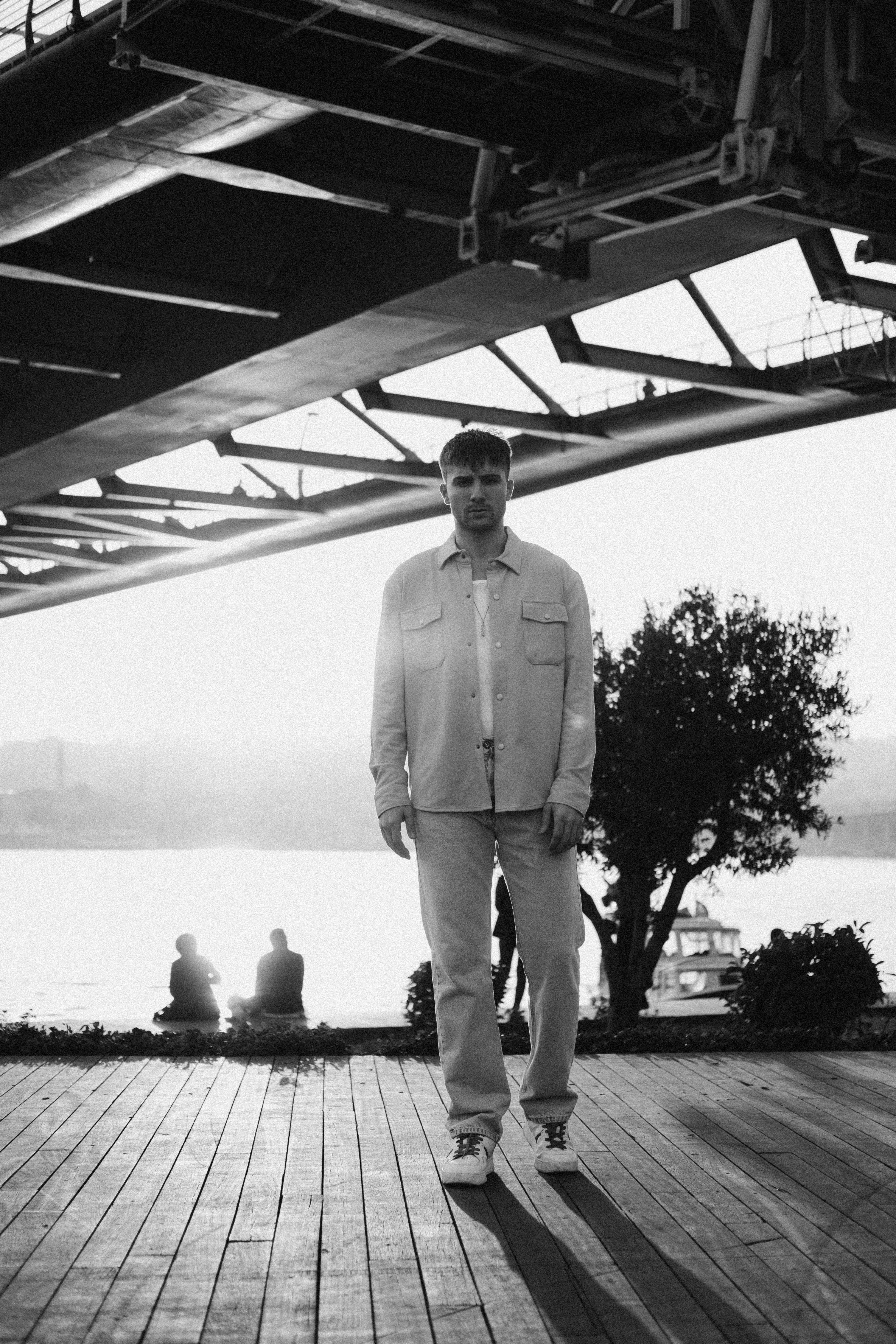Black and white photo of a man standing under a bridge by a riverside in Turkey.