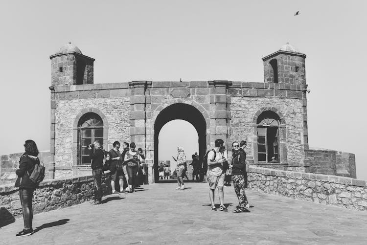 A Grayscale Photo Of People Standing Near Sqala Du Port D'Essaouira
