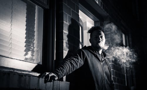 A Man in Black Jacket Smoking Near a Brick Wall and Glass Window