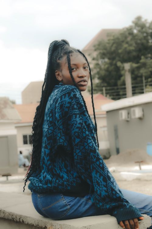 Free A fashionable woman with long braided hair sitting outdoors wearing a blue patterned sweater. Stock Photo