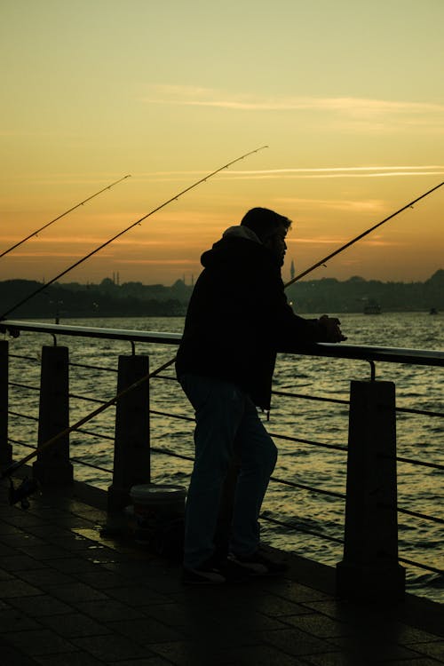 A Man Wearing a Black Jacket Standing on a Wooden Dock with Metal Railing with Fishing Rods
