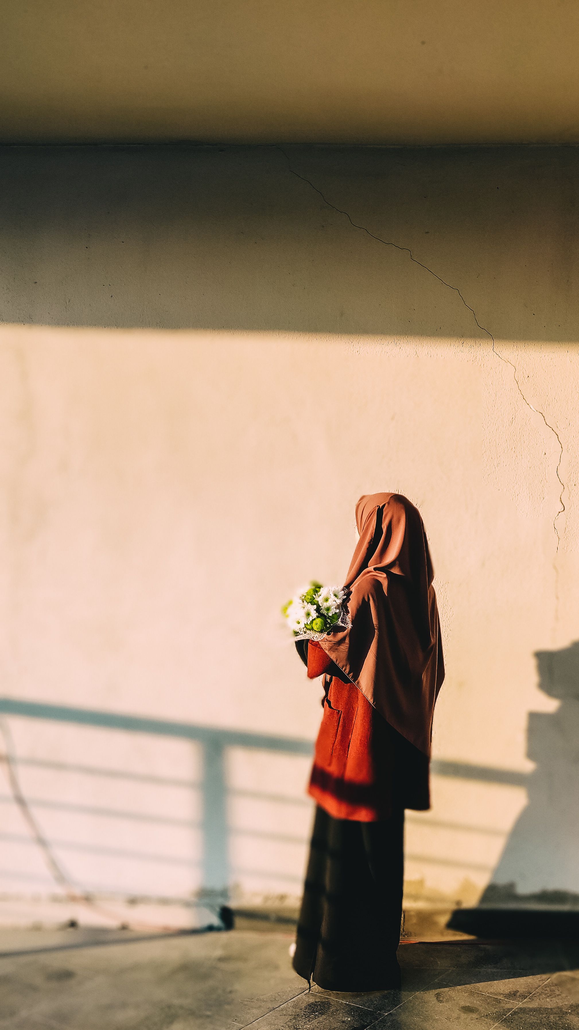 A woman in a hijab holds a flower bouquet, standing in sunlight, casting shadows.