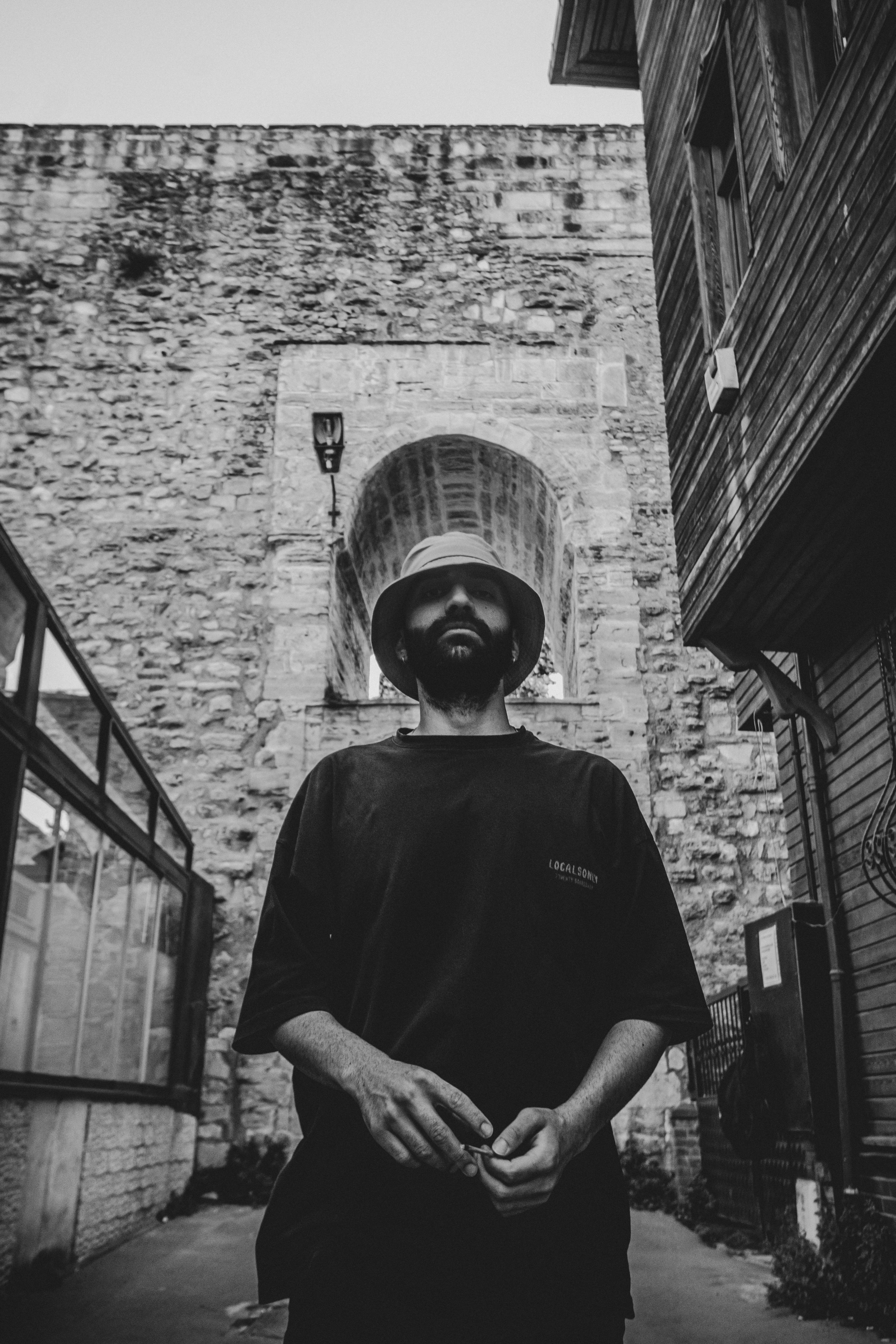 Grayscale portrait of a bearded man in cityscape with historic stone walls.