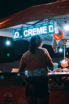 A woman serves customers at a bustling night market food stall under neon lights.
