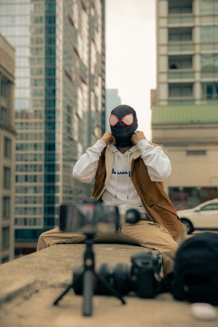 Man In A Mask Sitting At The Edge Of A Roof In Front Of A Phone