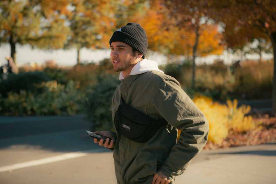 Young man wearing a green jacket and beanie, holding a smartphone in a park during fall.