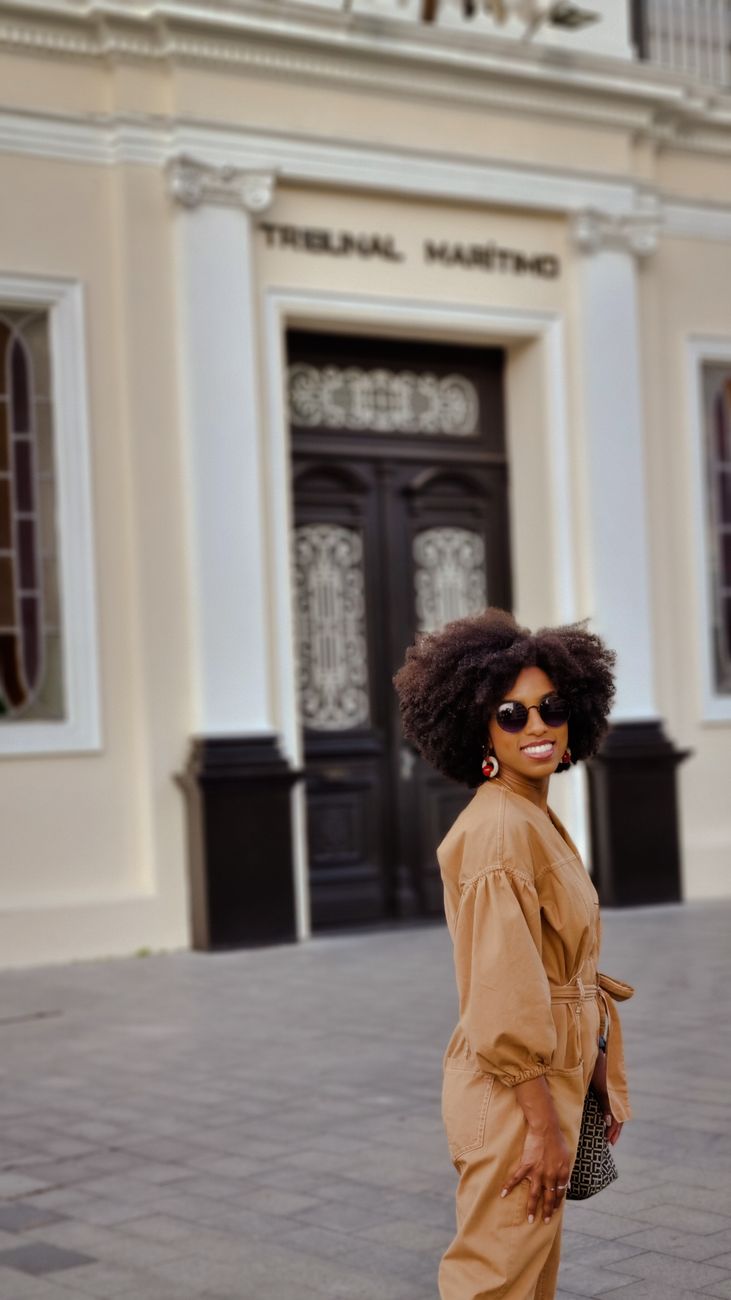 Mulher estilosa com cabelo afro no Rio de Janeiro, usando óculos de sol e um traje bege, em frente ao Tribunal Marítimo.