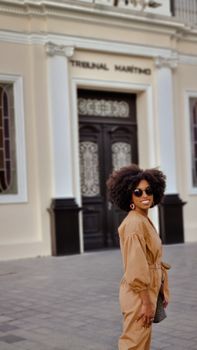 Stylish woman with afro hair in Rio de Janeiro, wearing sunglasses and a beige outfit, standing in front of Tribunal Marítimo.
