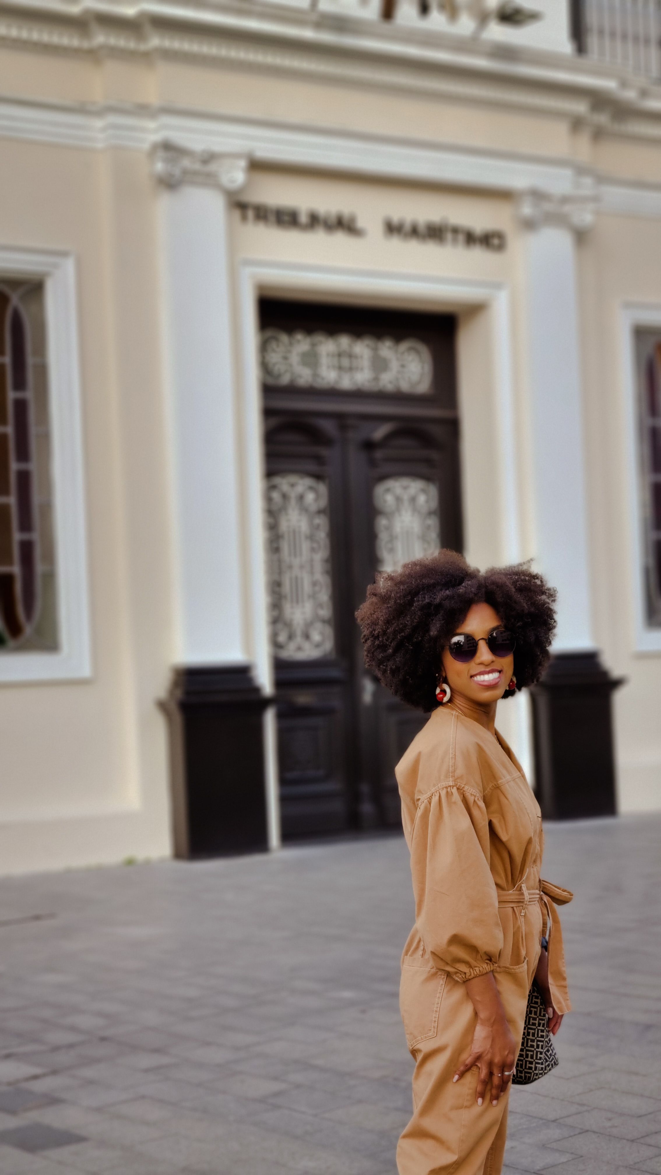 Mulher estilosa com cabelo afro no Rio de Janeiro, usando óculos de sol e um traje bege, em frente ao Tribunal Marítimo.