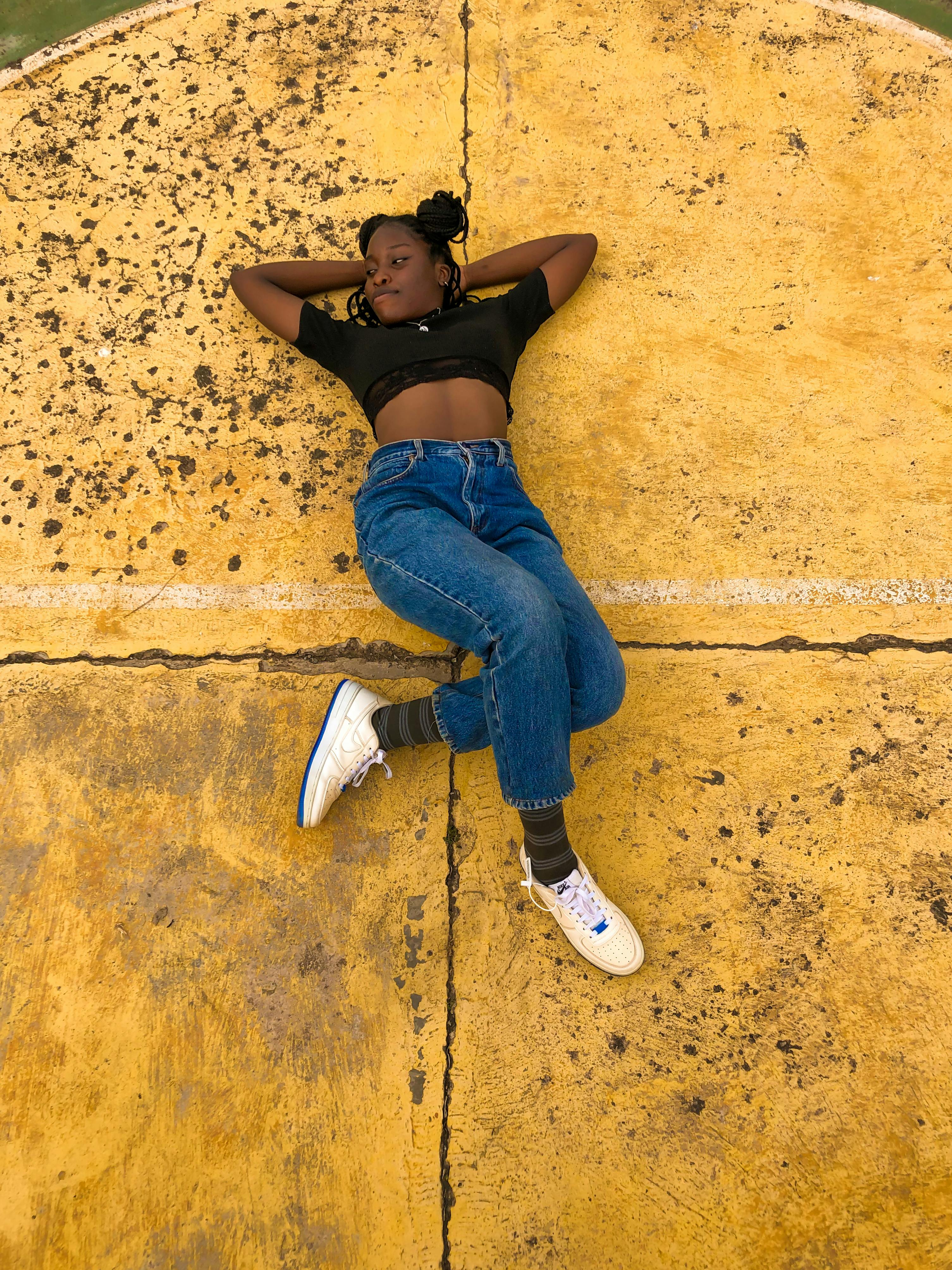 Young woman in crop top and denim pants lying on a vibrant yellow concrete surface, exuding a casual and relaxed vibe.