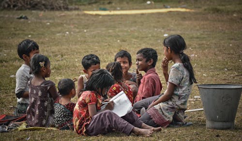 Group of full body ethnic children sitting on lawn and spending time together while unrecognizable little girl diligently doing homework wring in sheet of paper on street