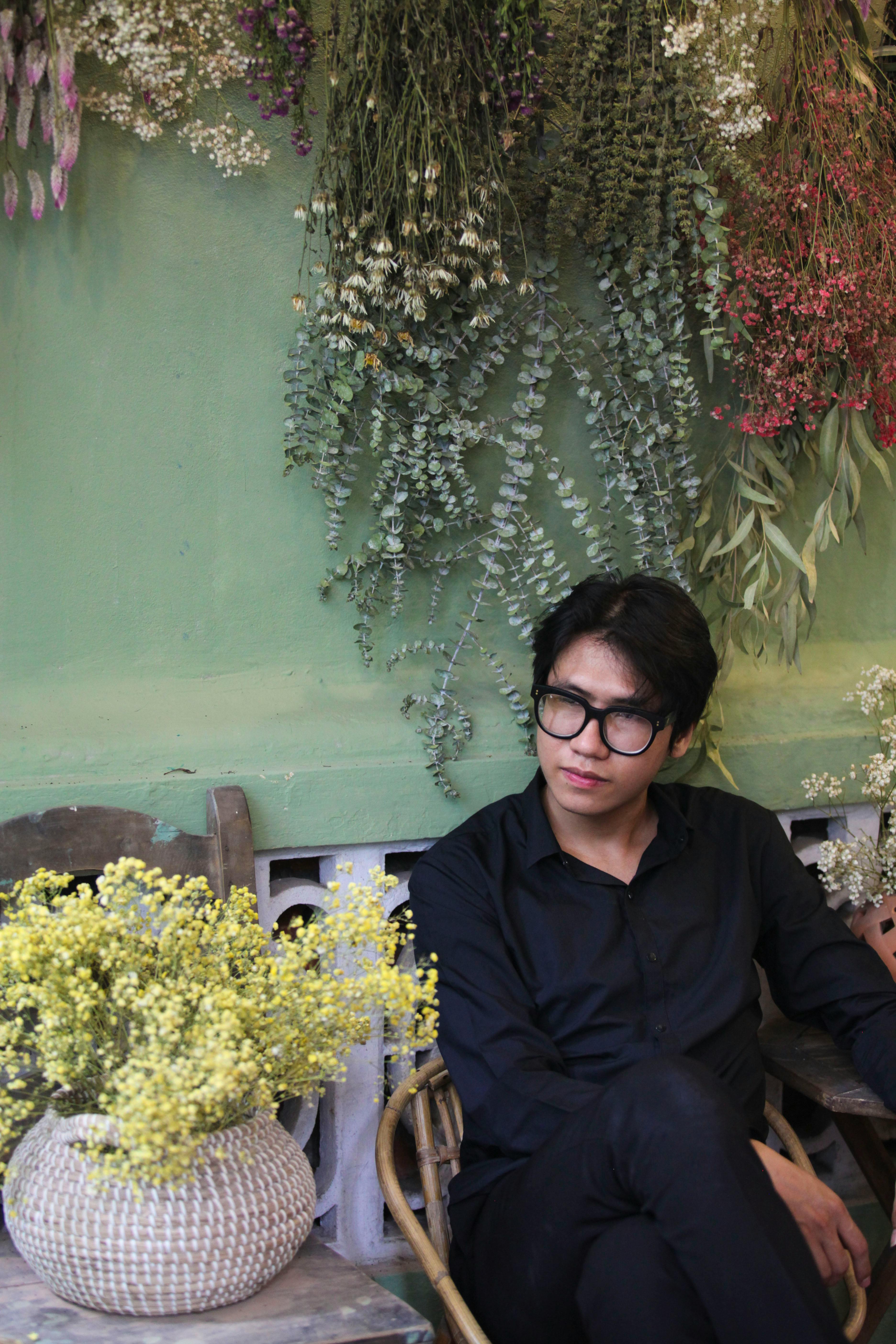 Man in black attire sitting indoors surrounded by floral decor, creating a serene ambiance.