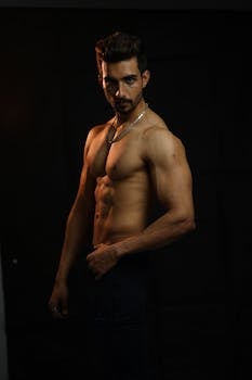 Muscular man posing shirtless in a studio setting, showcasing fitness and strength.