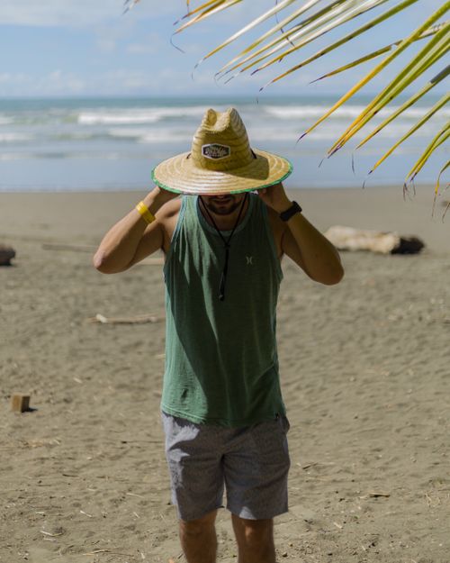 Free Man wearing a sun hat and gray shorts stands on a tropical beach under palm trees. Stock Photo
