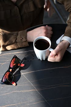 A person in brown attire holding a coffee cup on a table with sunglasses.