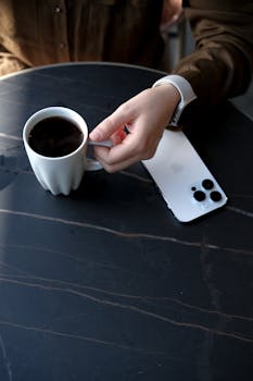 A person holding a cup of black coffee with a smartphone on the table, showcasing a casual moment.
