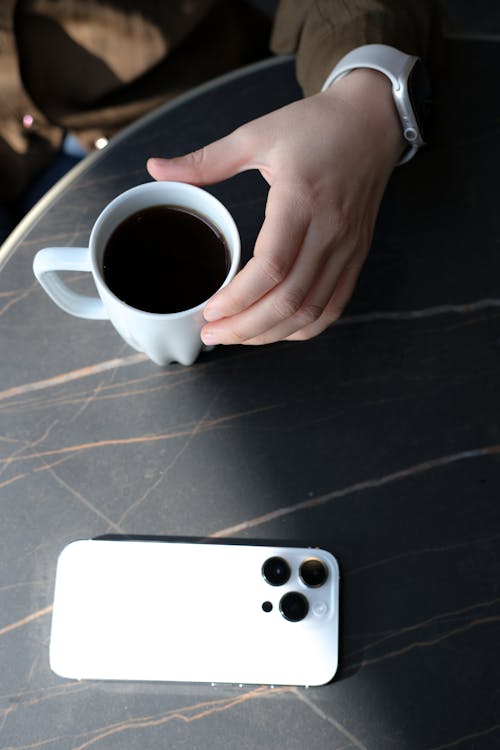 Person Holding a Cup of Coffee Beside a Smartphone