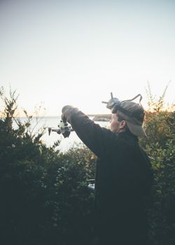 A person prepares to launch a drone near the sea at sunset, surrounded by bushes.