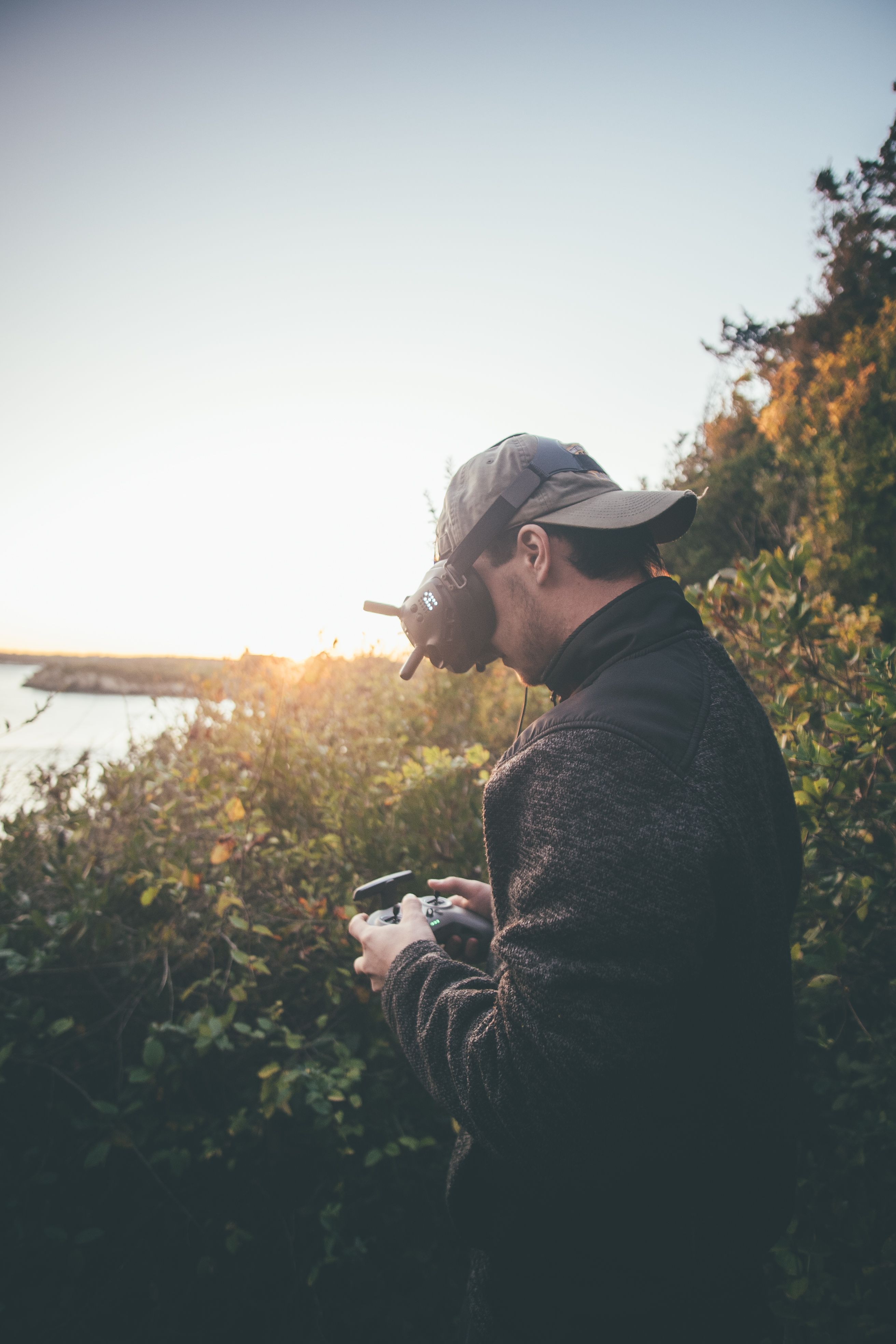 Adult man using VR goggles and a drone controller in a scenic outdoor setting during sunset.
