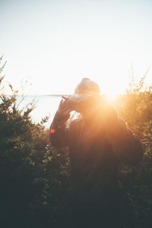 Free A person stands outdoors wearing virtual reality goggles against a sunset backdrop. Stock Photo
