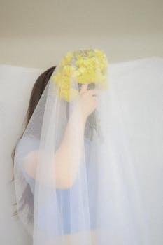 A woman holds yellow flowers under a delicate wedding veil, creating a soft, artistic look.