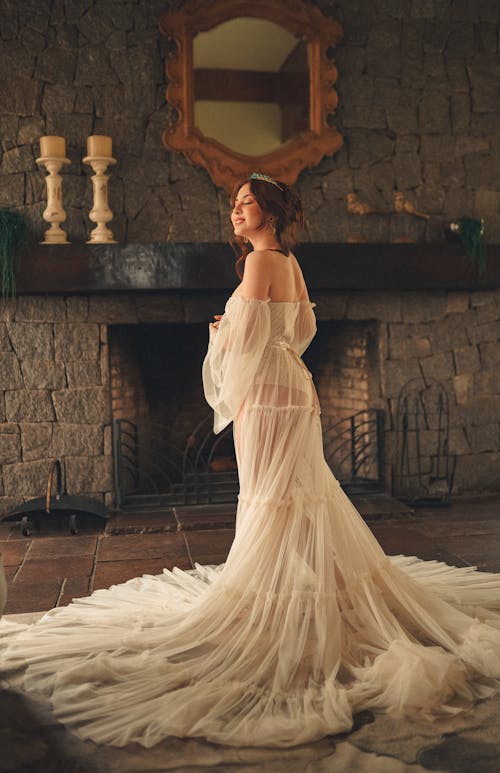 Woman in White Long Dress Standing Beside a Fireplace