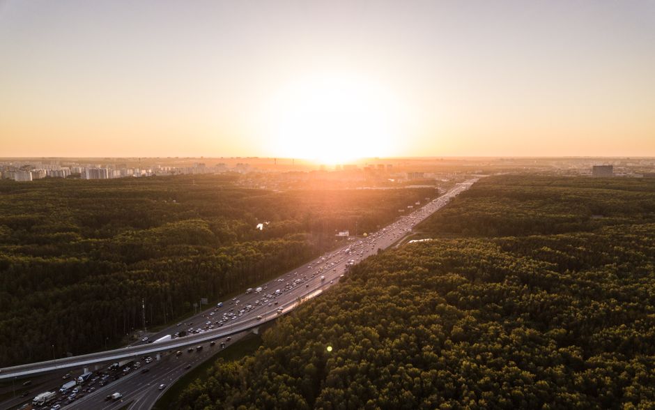A scenic aerial view of a highway cutting through a dense forest at sunset with cityscape in the background.