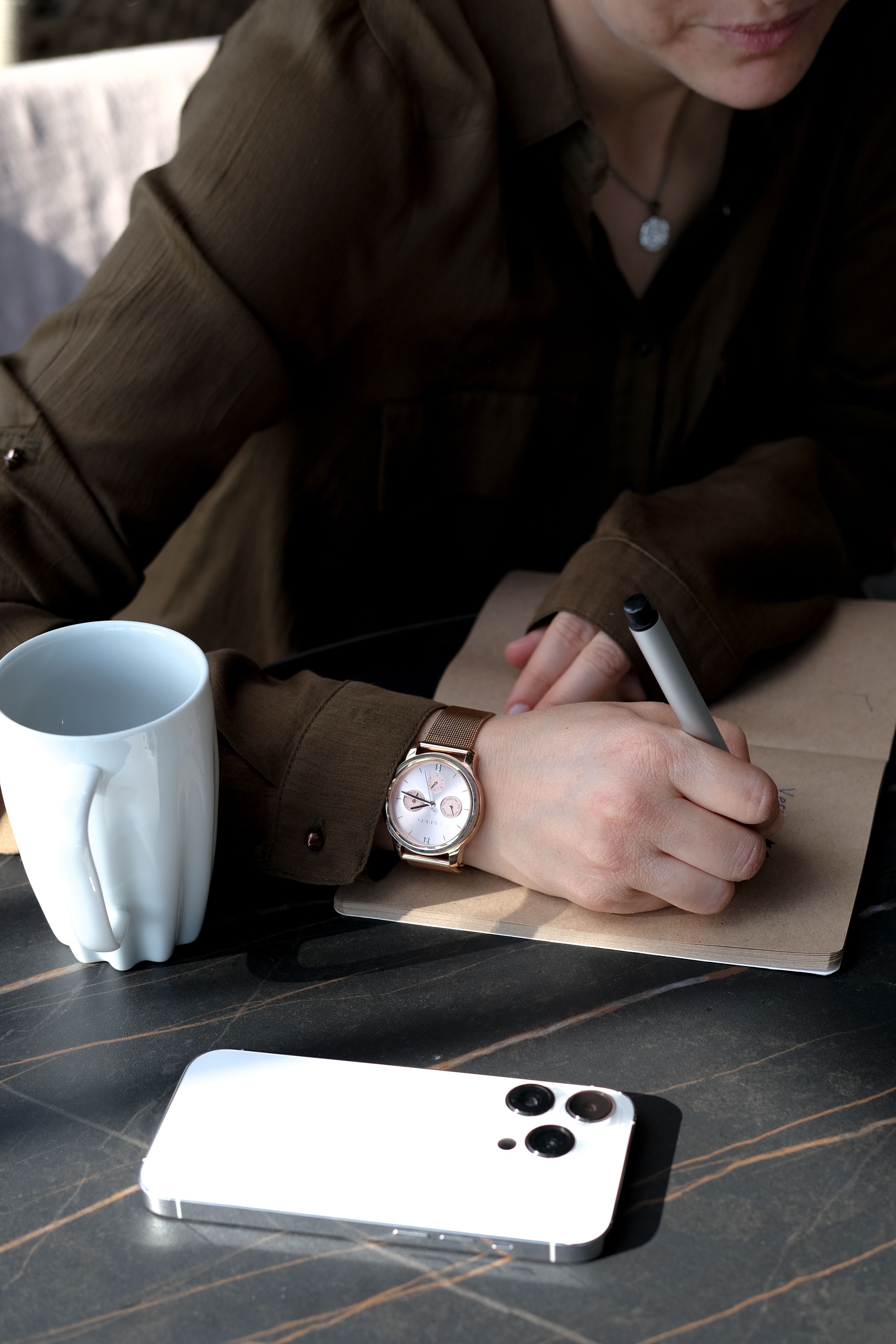 A woman writing in a notebook at a table beside a smartphone and a white cup