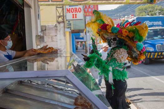 A colorful Dia de los Muertos character in costume buys pastries on a bustling street.